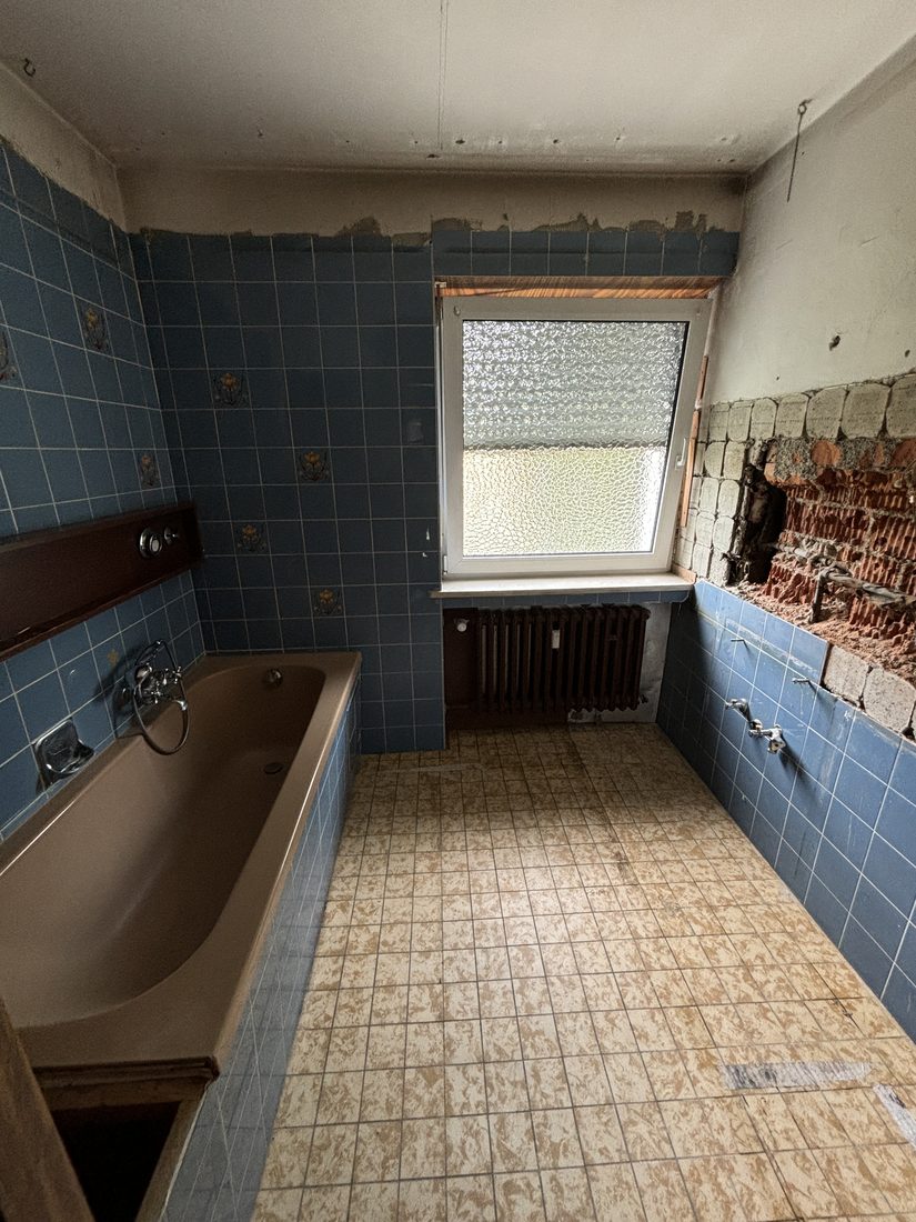 Bad in Dachau vor der Sanierung: blaue Fliesen, alte Wanne, teilweise abgeschlagene Wand.