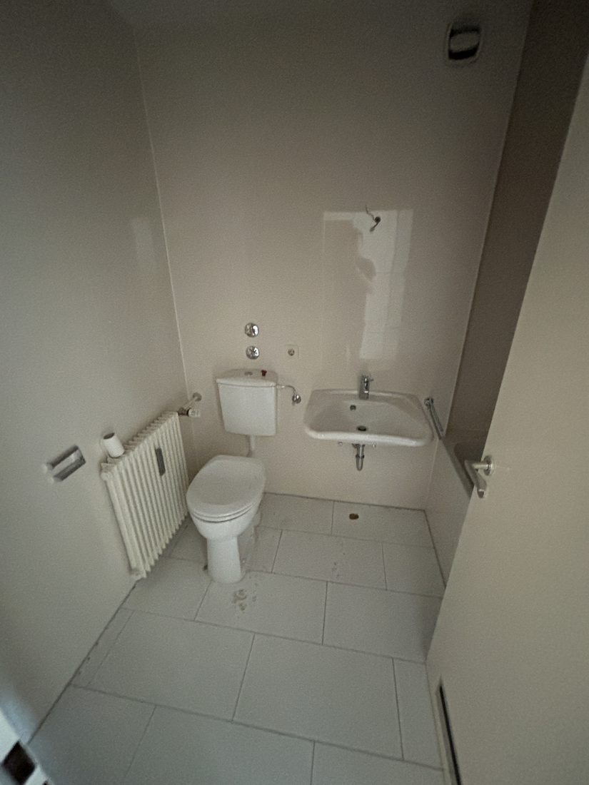 Bad in München-Sendling vor der Sanierung: schmaler weiß gefliester Raum mit altem Spülkasten-WC, kleinem Waschbecken und Rippenheizkörper.